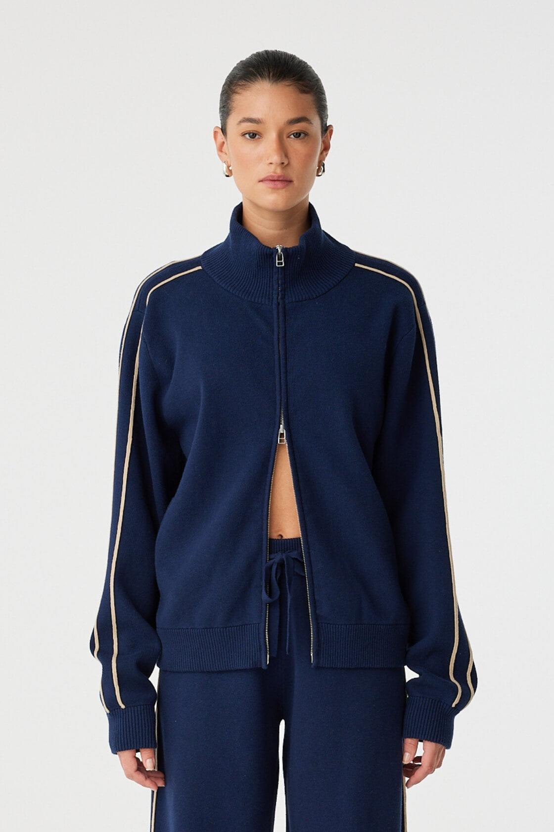 Model wears navy track-style zip jacket with ribbed funnel neck and contrast sleeve stripes.
