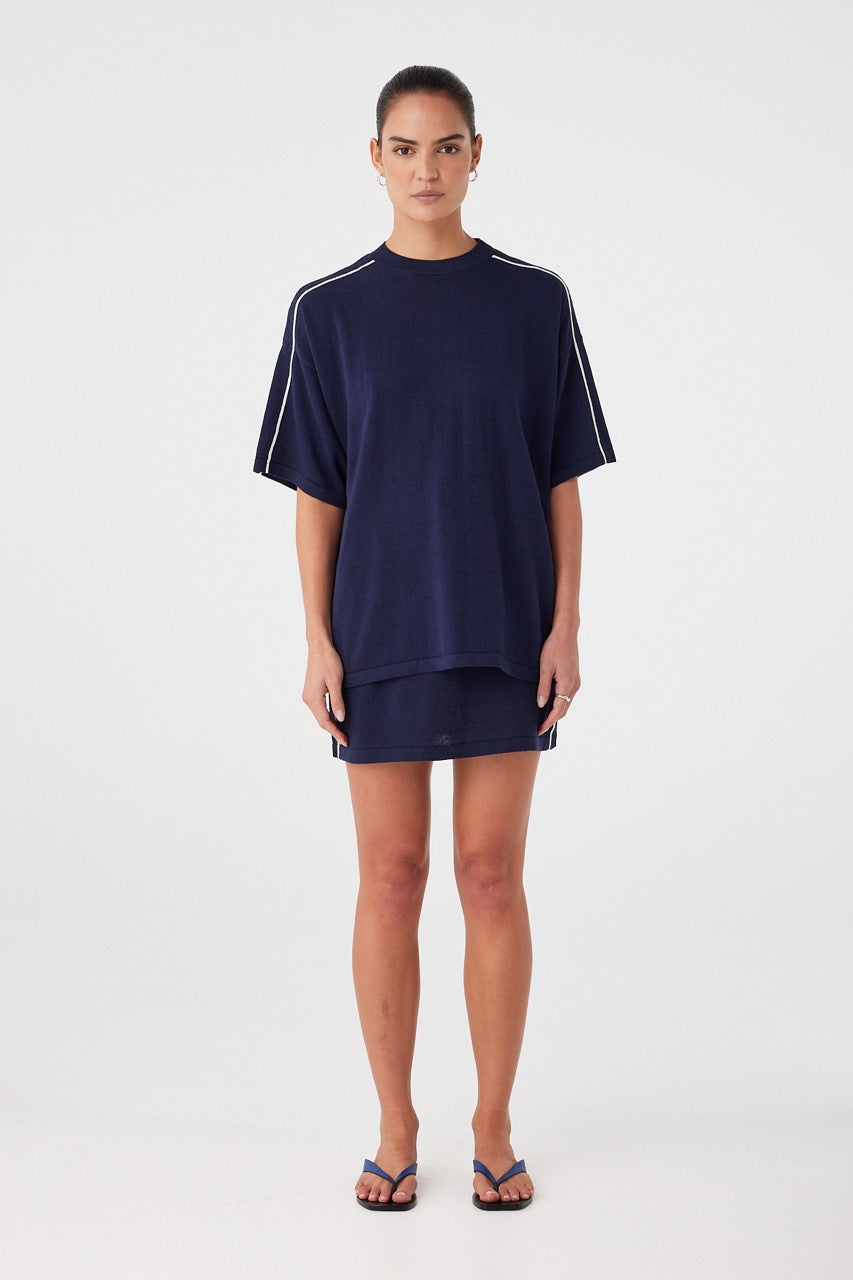 Model wears navy mini skirt in linen–cotton knit with contrast white side stripes. Straight silhouette with ribbed waistband and drawcord.