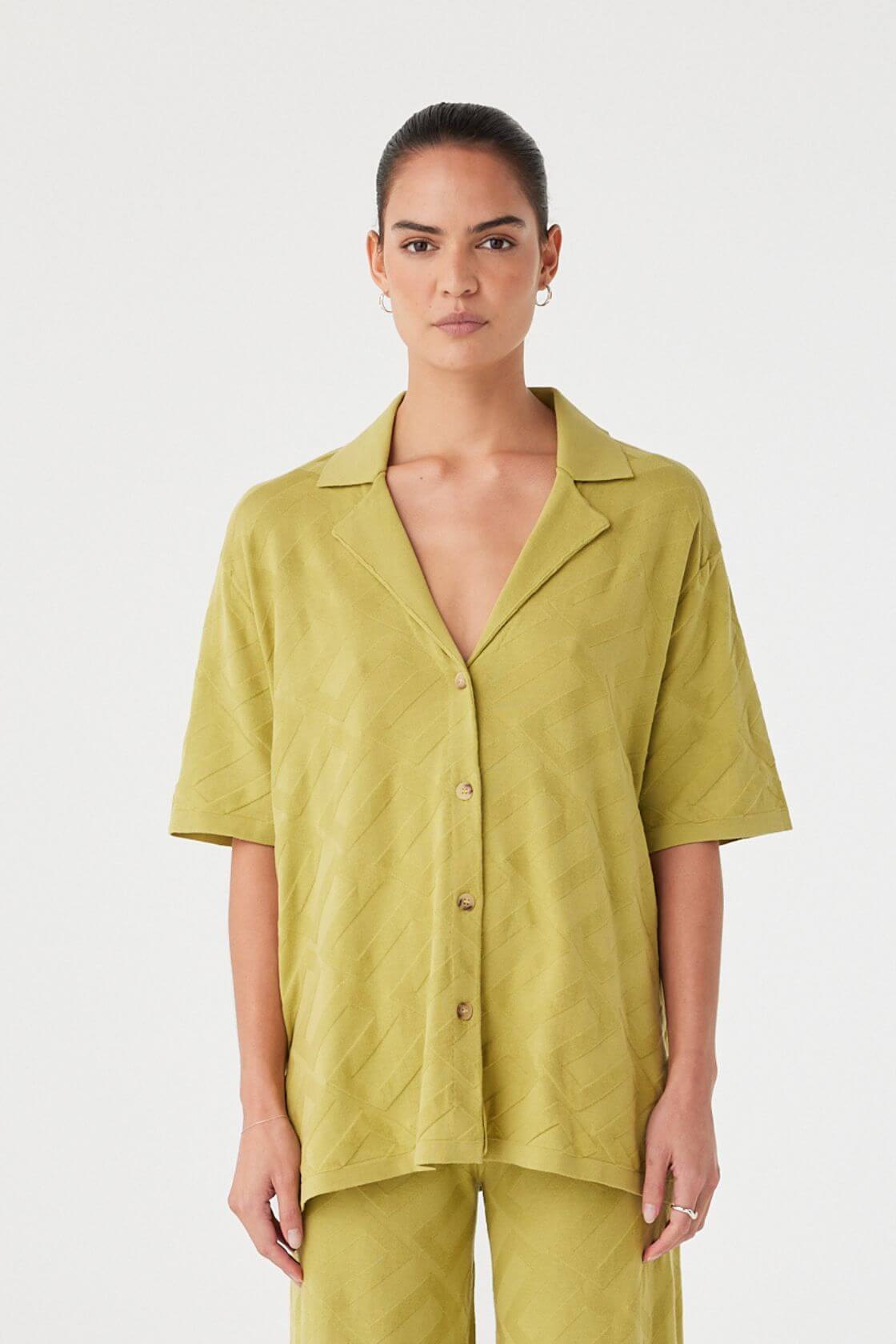 Model wears an oversized short-sleeve shirt in matcha green organic cotton knit. Breathable texture with button front and open collar.