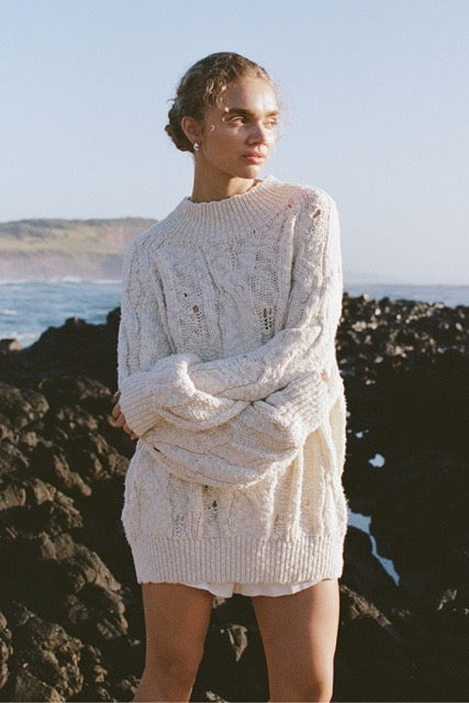 Lifestyle image of cream chunky cable knit sweater worn outdoors by the ocean