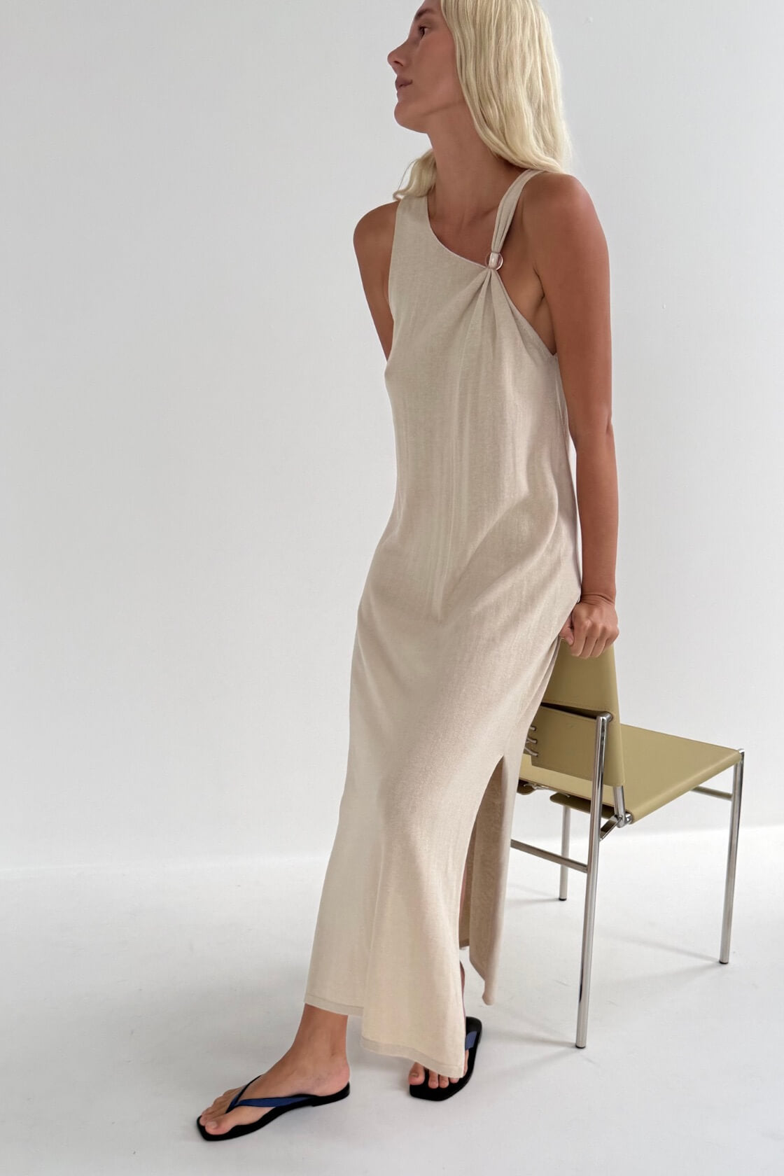 A woman modeling a sleeveless, taupe-colored linen blend maxi dress with a high side slit, standing next to a minimalist beige chair against a white studio background.