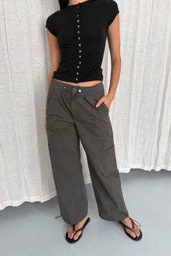 Model wearing the Addie pants in Charcoal, a black short-sleeve button-front top, and black thong sandals, standing in front of a white curtain backdrop.