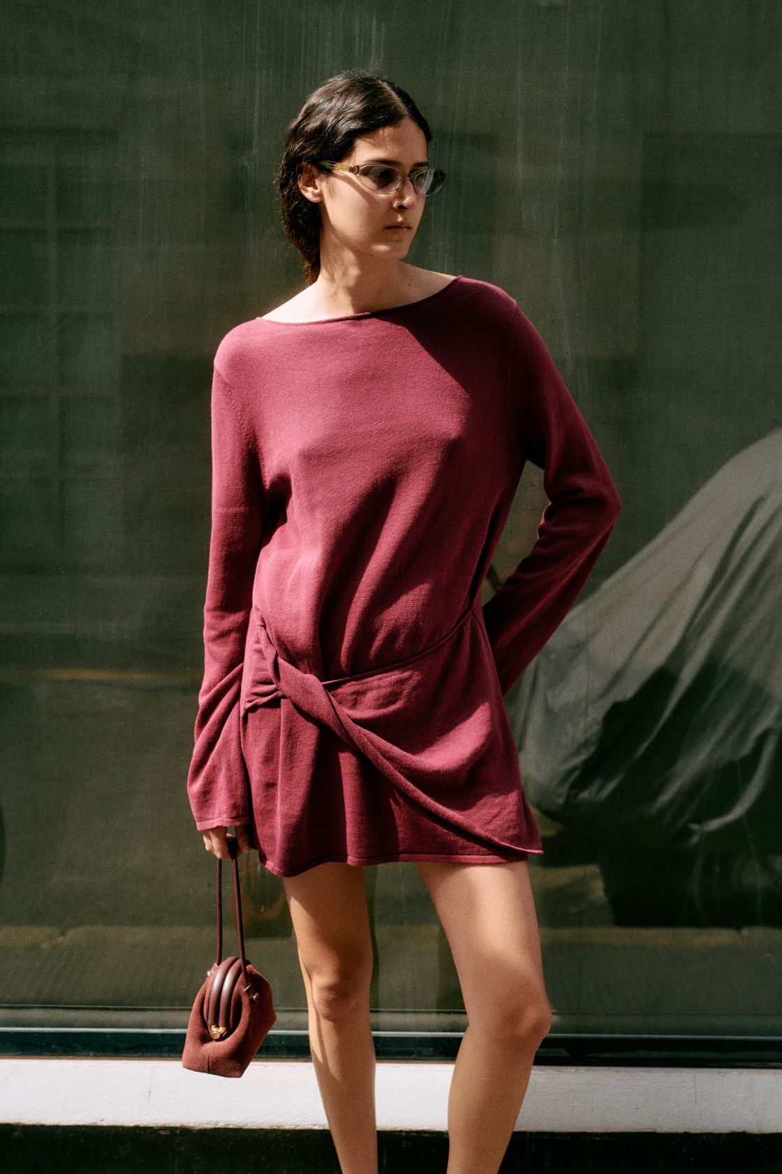 Model wearing the Kira mini dress in merlot against a green background with a maroon bag. 