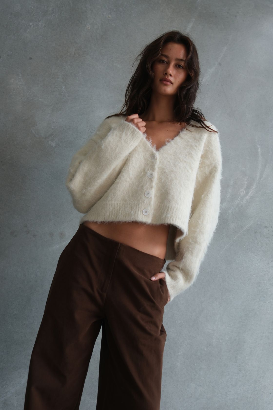 Model wearing the Liora Fluffy Cardigan in Cream, featuring a soft brushed knit texture, cropped silhouette and button-front design, styled with espresso wide-leg trousers against a textured concrete backdrop.