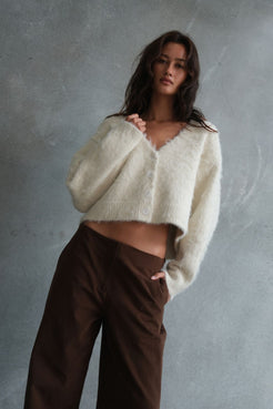 Model wearing the Liora Fluffy Cardigan in Cream, featuring a soft brushed knit texture, cropped silhouette and button-front design, styled with espresso wide-leg trousers against a textured concrete backdrop.
