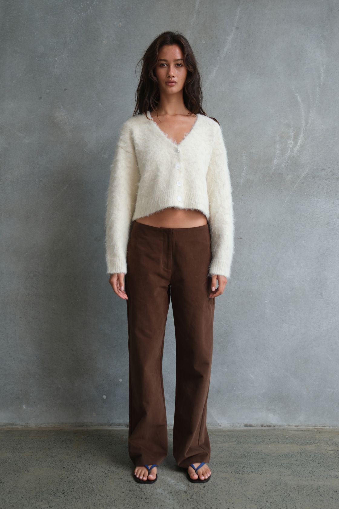 Model wearing the Avery Pant in Espresso, showcasing the full-length straight-leg fit and rich espresso tone, paired with a cream fluffy cropped cardigan against a textured concrete backdrop.
