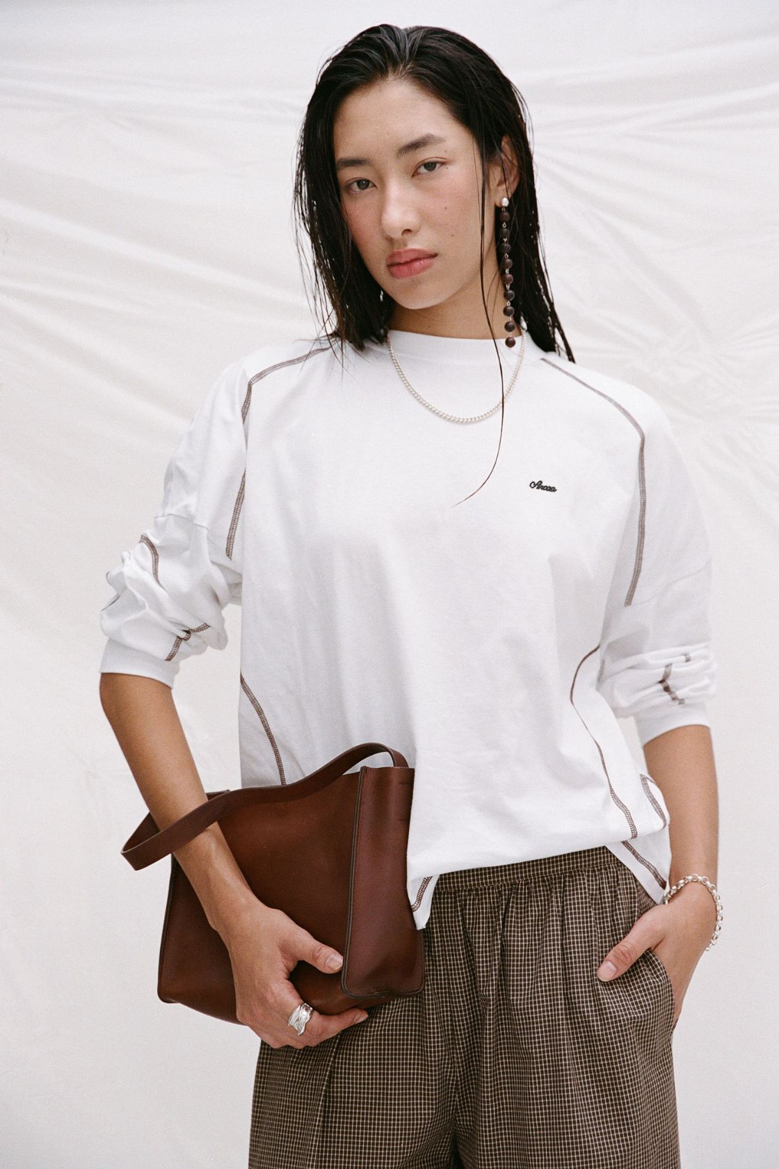 Model wearing the Kesh tee in cream with chocolate check pants and a brown leather bag. 