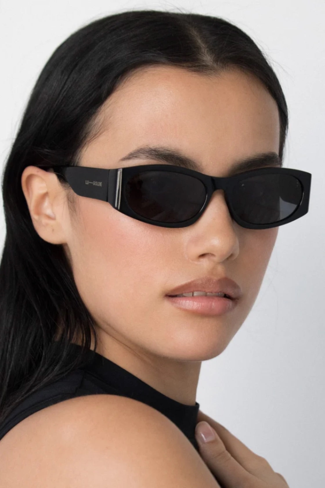 A close-up of a model wearing black sporty cat-eye sunglasses with a thin silver metal accent on the temple arm, set against a plain light gray background.