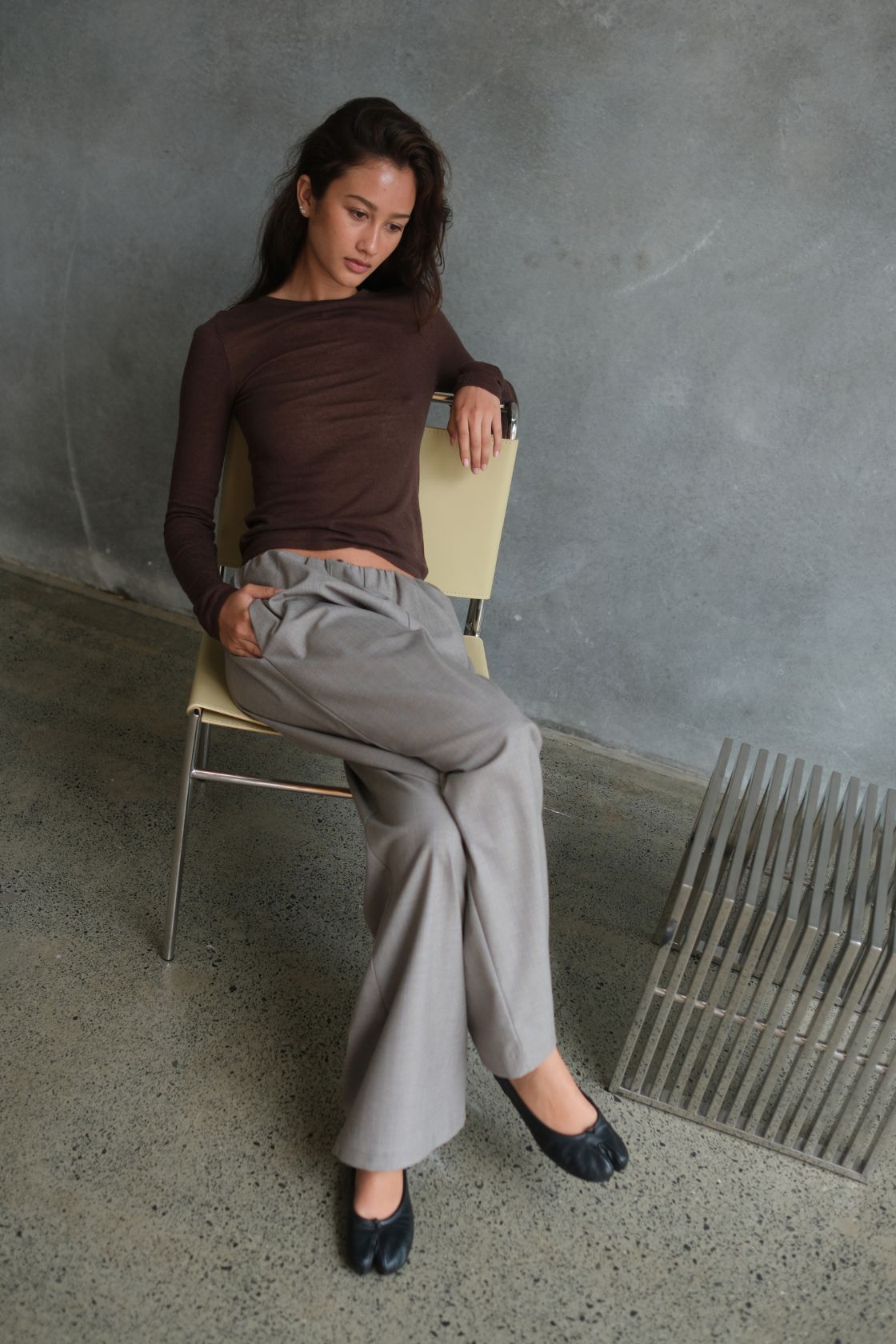Model wearing the Annika Pant in Taupe & Blue Pinstripe, featuring a relaxed wide-leg silhouette with subtle vertical pinstripes, styled with a fitted chocolate brown long-sleeve top while seated against a textured concrete backdrop.