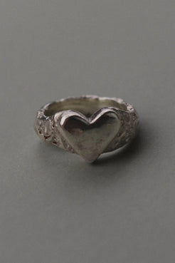 A textured sterling silver ring featuring a raised, polished heart-shaped centerpiece, photographed on a smooth, neutral gray background.