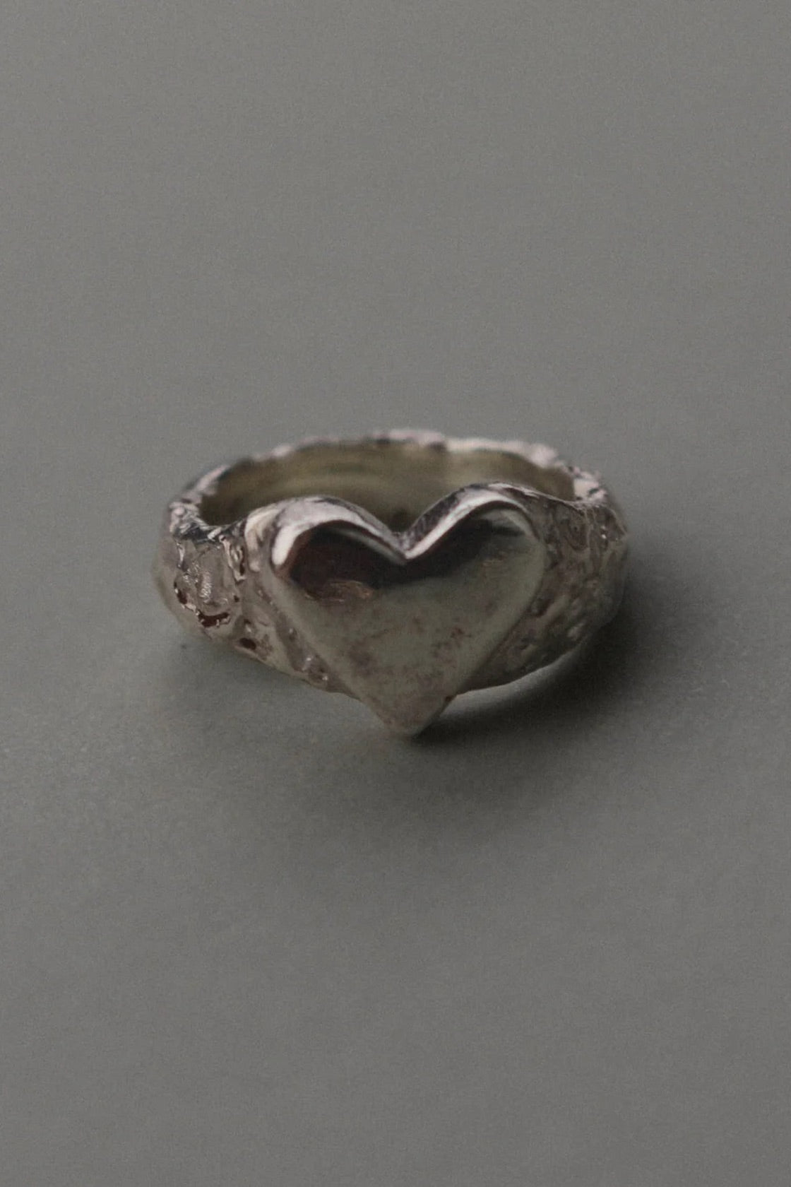 A textured sterling silver ring featuring a raised, polished heart-shaped centerpiece, photographed on a smooth, neutral gray background.