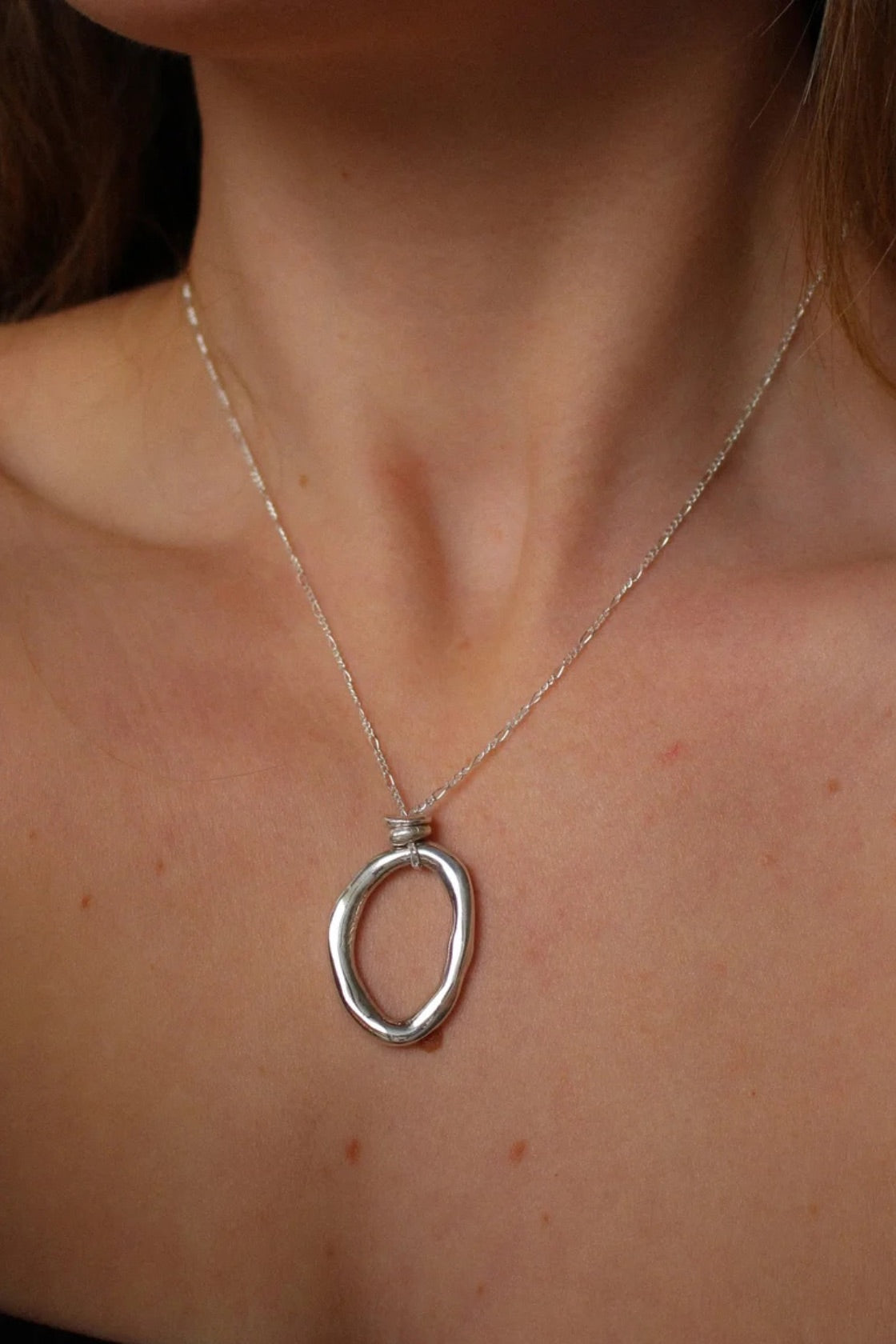 A close-up of a person's neck and chest wearing a silver chain necklace with a polished, open oval pendant.