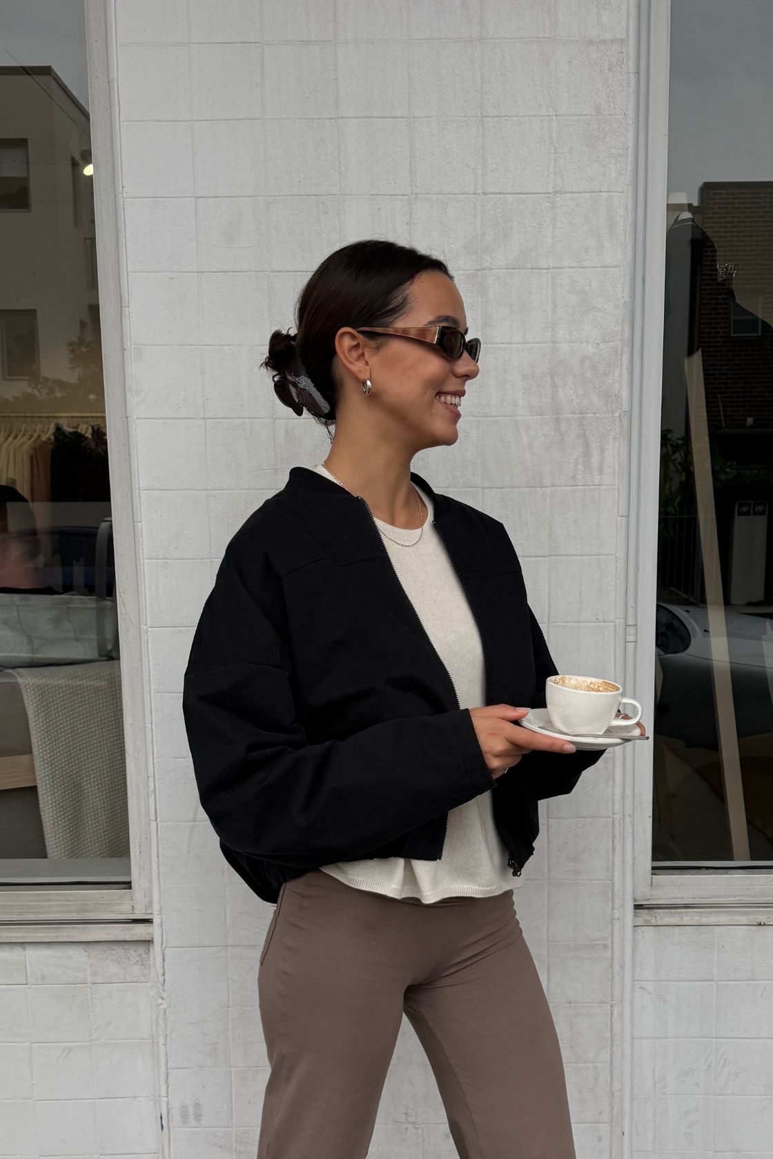 Lifestyle image of a model wearing the Wren Bomber in black featuring a relaxed oversized fit, cropped silhouette and zip-front closure, styled with taupe cropped pants and a cream knit top while holding a coffee outside a cafe