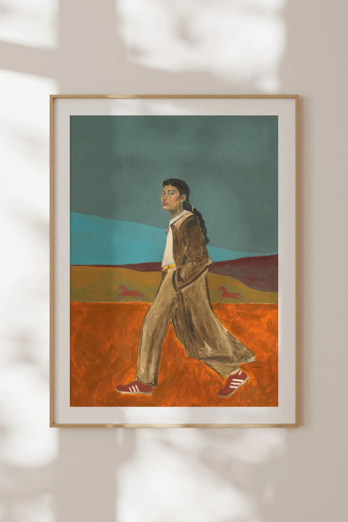 A framed art print featuring a stylized illustration of a woman walking across a landscape, displayed on a light beige wall with soft leaf shadows.