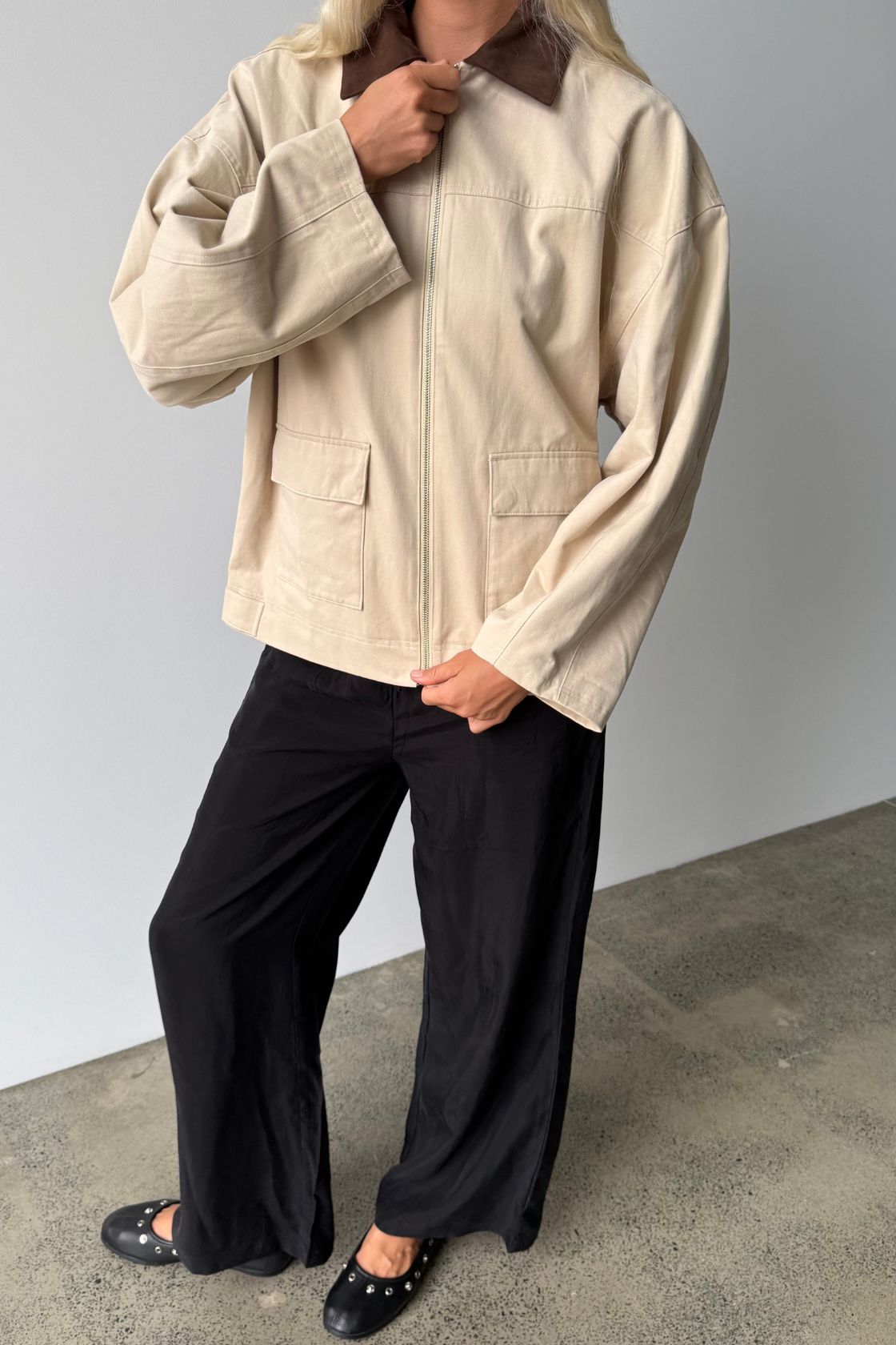 Model wearing a cream cotton jacket with an espresso contrast collar, oversized sleeves and front zip closure, styled with relaxed black wide-leg pants and black studded flats.