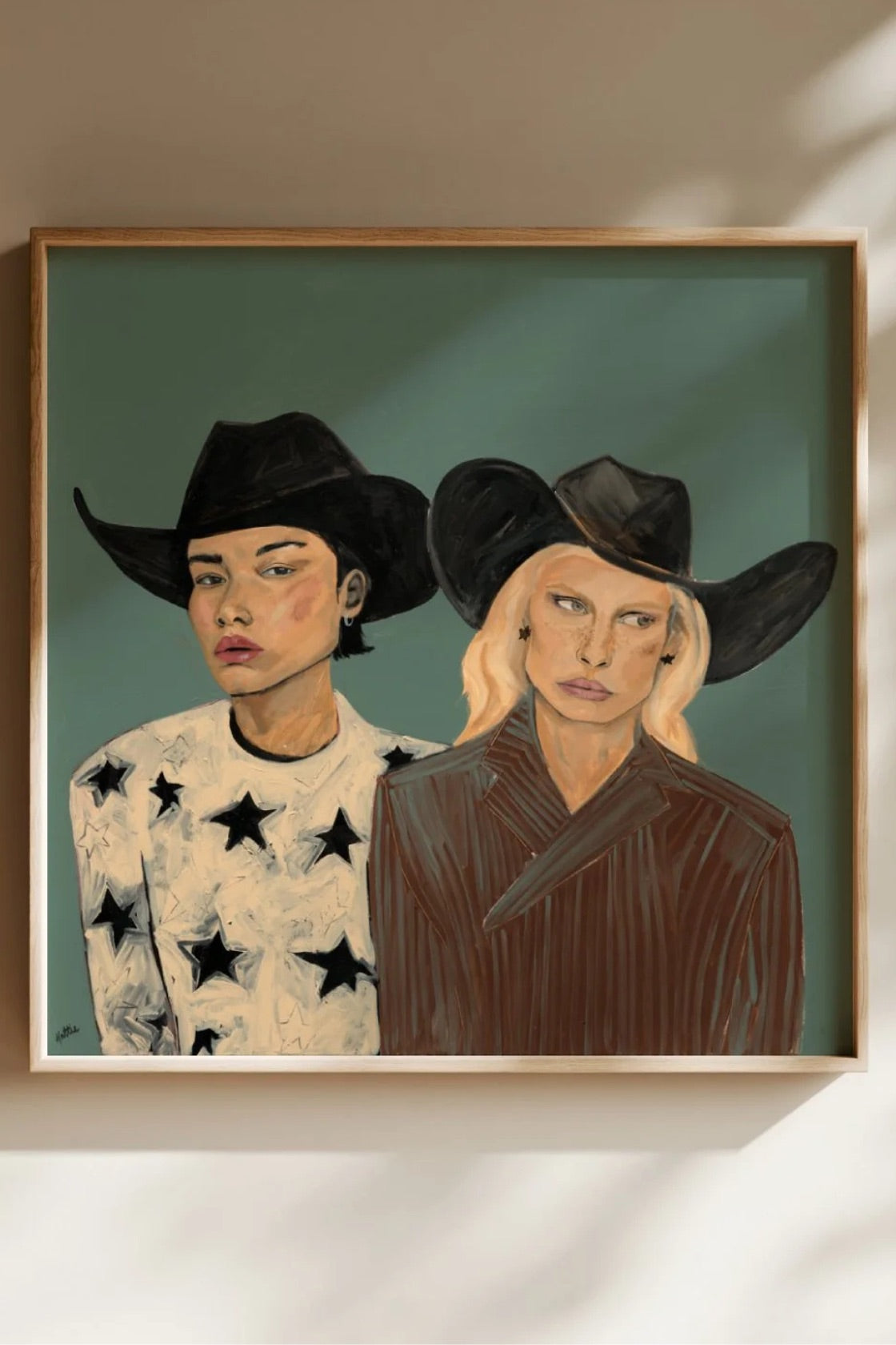 A framed portrait painting of two women in cowboy hats, displayed on a beige wall with soft shadows. The left figure wears a white shirt with black stars, and the right figure wears a dark brown top.
