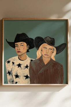 A framed portrait painting of two women in cowboy hats, displayed on a beige wall with soft shadows. The left figure wears a white shirt with black stars, and the right figure wears a dark brown top.