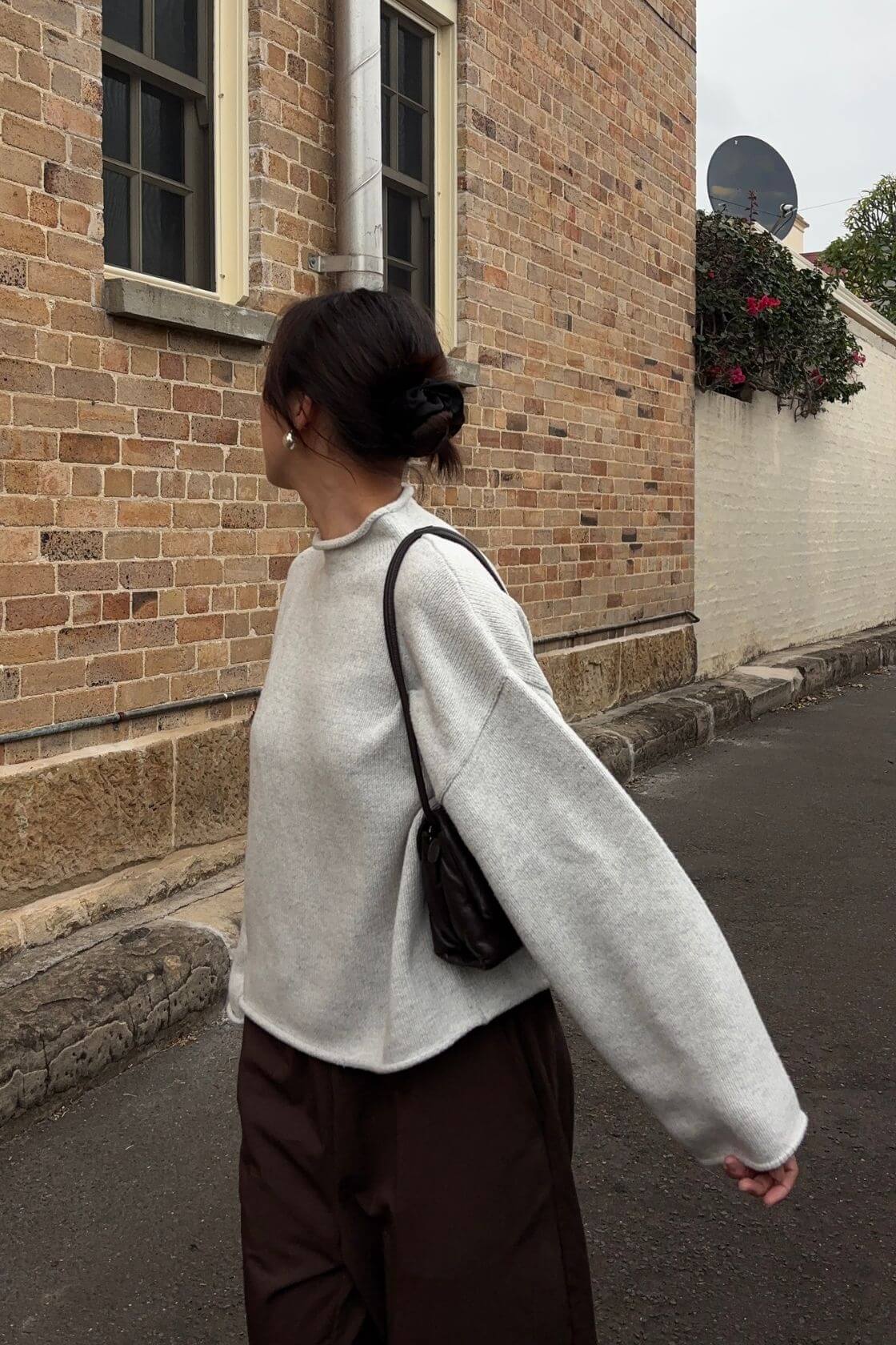 Lifestyle side view of the Maeve Sweater in snow marle featuring a soft lightweight knit texture, relaxed oversized silhouette and rolled neckline styling paired with espresso wide-leg pants and a black shoulder bag