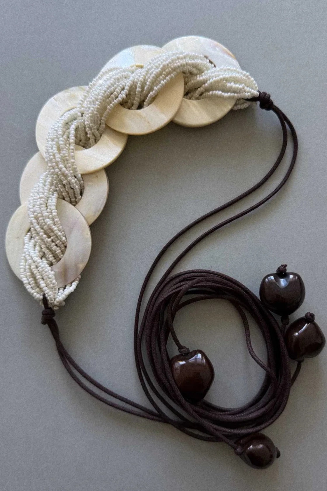 A multi-strand beaded necklace featuring cream-colored shell disc links and dark brown cords with wooden bead accents, displayed on a neutral gray background.
