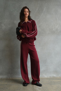 Model wearing the Nella Zip in Merlot & Cream, featuring a relaxed fit with contrast cream piping along the collar and sleeves, styled with matching wide-leg pants and a black pouch bag against a textured concrete backdrop.