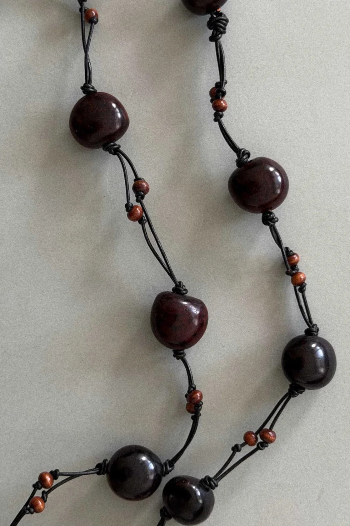 A close-up of a beaded belt featuring large, polished dark reddish-brown beads and small orange-red accent beads on a black cord, laid flat on a neutral light gray background.