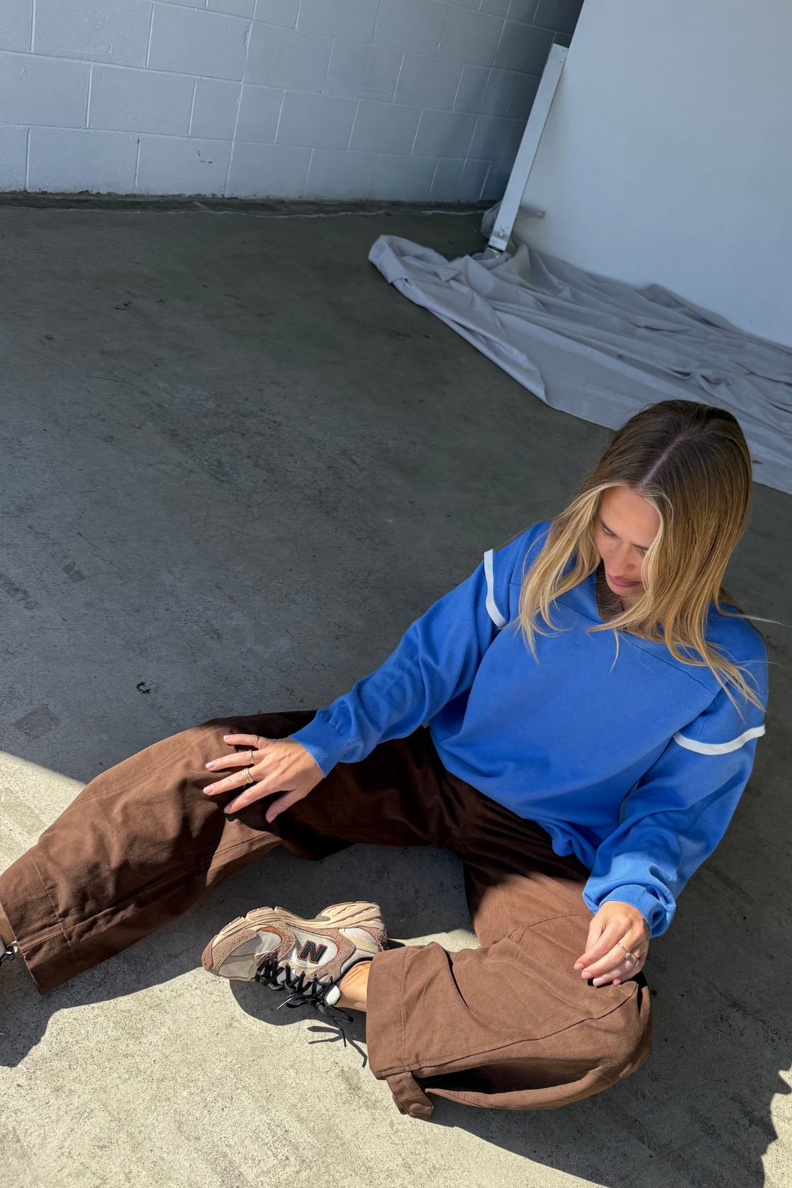 Lifestyle image of a model sitting on a concrete studio floor wearing a bright blue oversized sweatshirt with contrast stripe detailing on the sleeves, styled with chocolate brown wide-leg pants and neutral sneakers in natural light