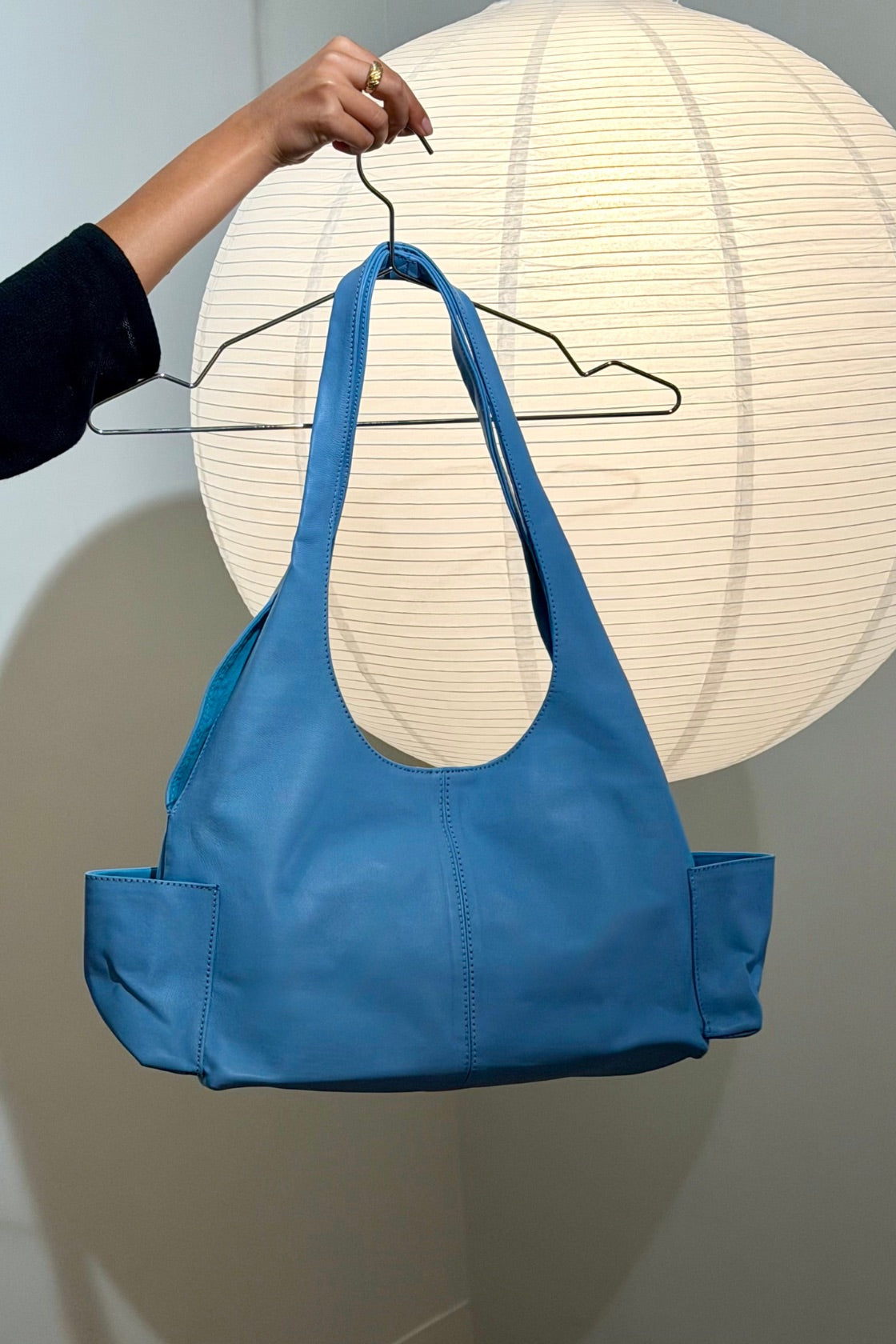 A bright blue leather shoulder bag with side pockets, hanging from a black wire hanger held by a person's hand in front of a large white paper lantern.