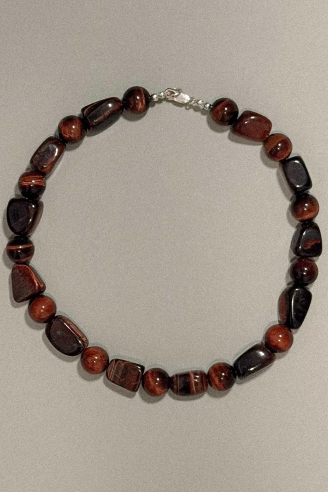 A beaded necklace made of polished reddish-brown and black gemstone beads with a silver clasp, displayed on a neutral light gray background.