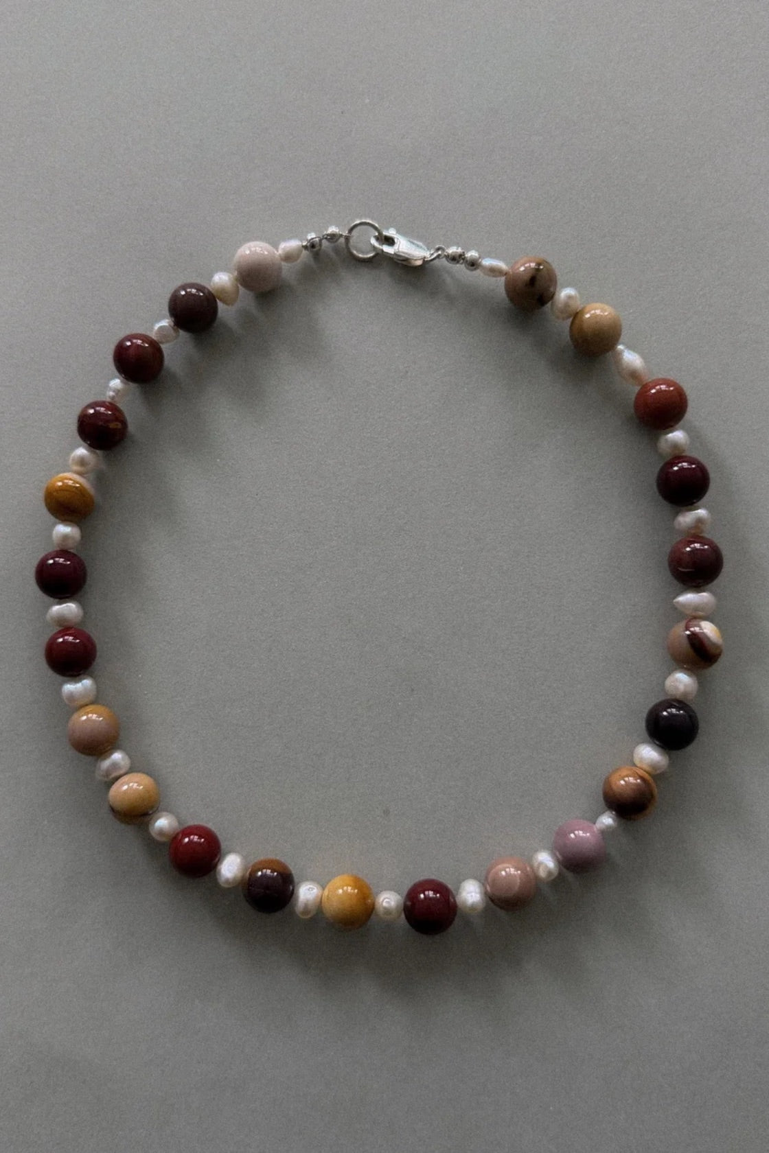 A beaded necklace with alternating polished gemstone-like round beads in dark red, brown, orange, and white, accented by small white pearl-like beads and a silver lobster clasp, displayed on a neutral gray background.