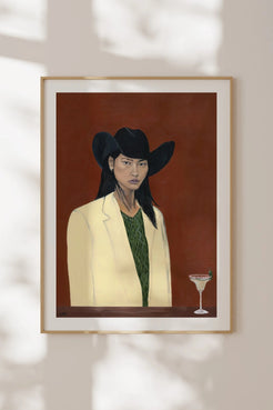 A framed art print featuring a stylized portrait of a woman in a black cowboy hat, cream blazer, and green top against a dark reddish-brown background, hanging on a light beige wall with soft shadow overlays.