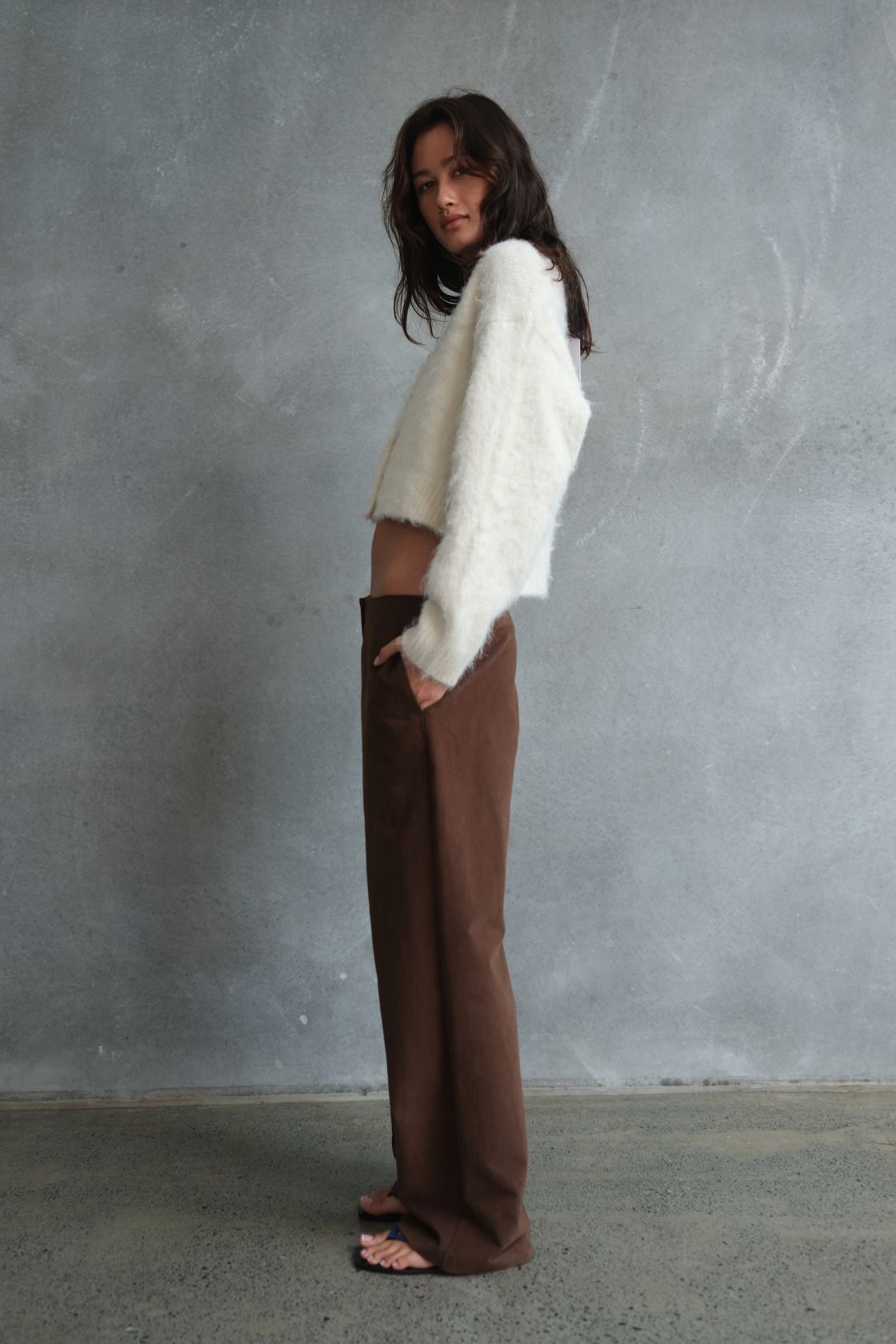Model wearing the Avery Pant in Espresso, featuring a relaxed straight-leg silhouette with a clean tailored finish, styled with a cream fluffy cropped cardigan and blue flip flops in a side profile against a textured concrete backdrop.