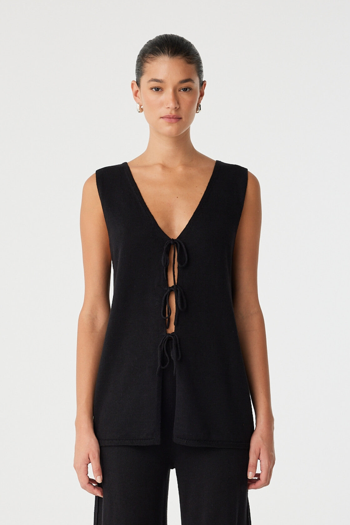 Model wears longline sleeveless top in black linen–cotton with deep V neckline and tie-front detail—styled for warm-weather ease.