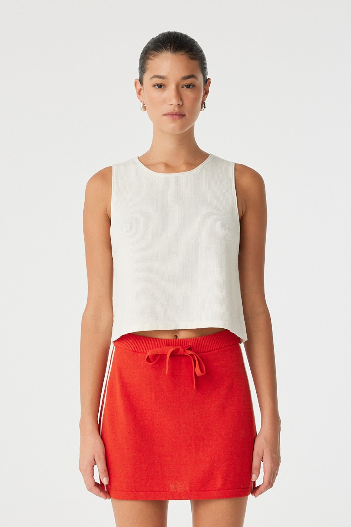 Model wears cream knit tank in lightweight cotton–hemp blend with rolled edges and relaxed flared shape—styled with a red mini skirt.