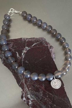 Taupe grey agate necklace with sterling silver Eddy Swirl pendant and clasp, draped over a dark stone surface.