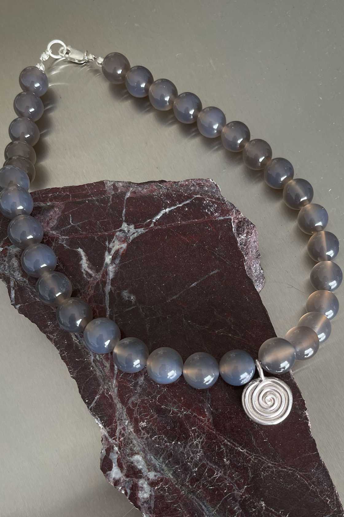 Taupe grey agate necklace with sterling silver Eddy Swirl pendant and clasp, draped over a dark stone surface.