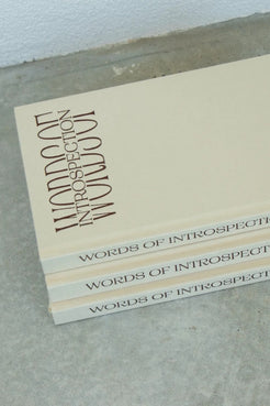 Three copies of the book 'Words of Introspection" stacked on a gray concrete surface against a white wall.