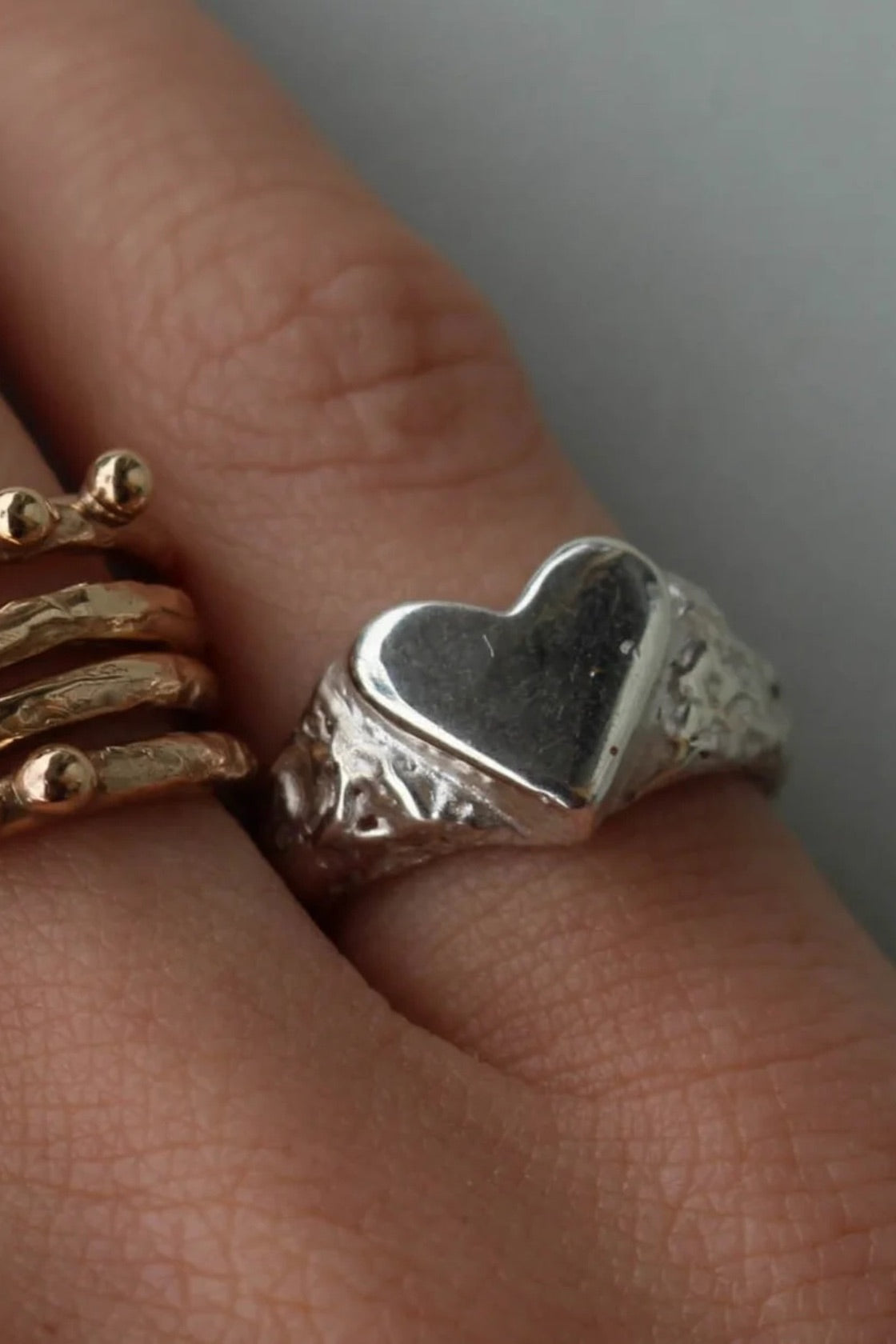 A close-up of a hand wearing a silver signet-style ring with a heart-shaped face and textured band, alongside stacked gold rings on adjacent fingers.
