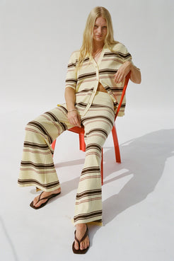A woman sits on a bright orange chair wearing a butter-colored two-piece matching set with brown and black horizontal stripes. The set includes a short-sleeve button-up shirt and wide-leg pants.