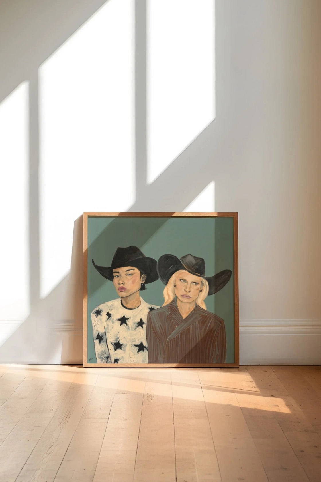 A framed art print depicting two women in cowboy hats, leaning against a white wall on a light wood floor with dramatic window shadows.