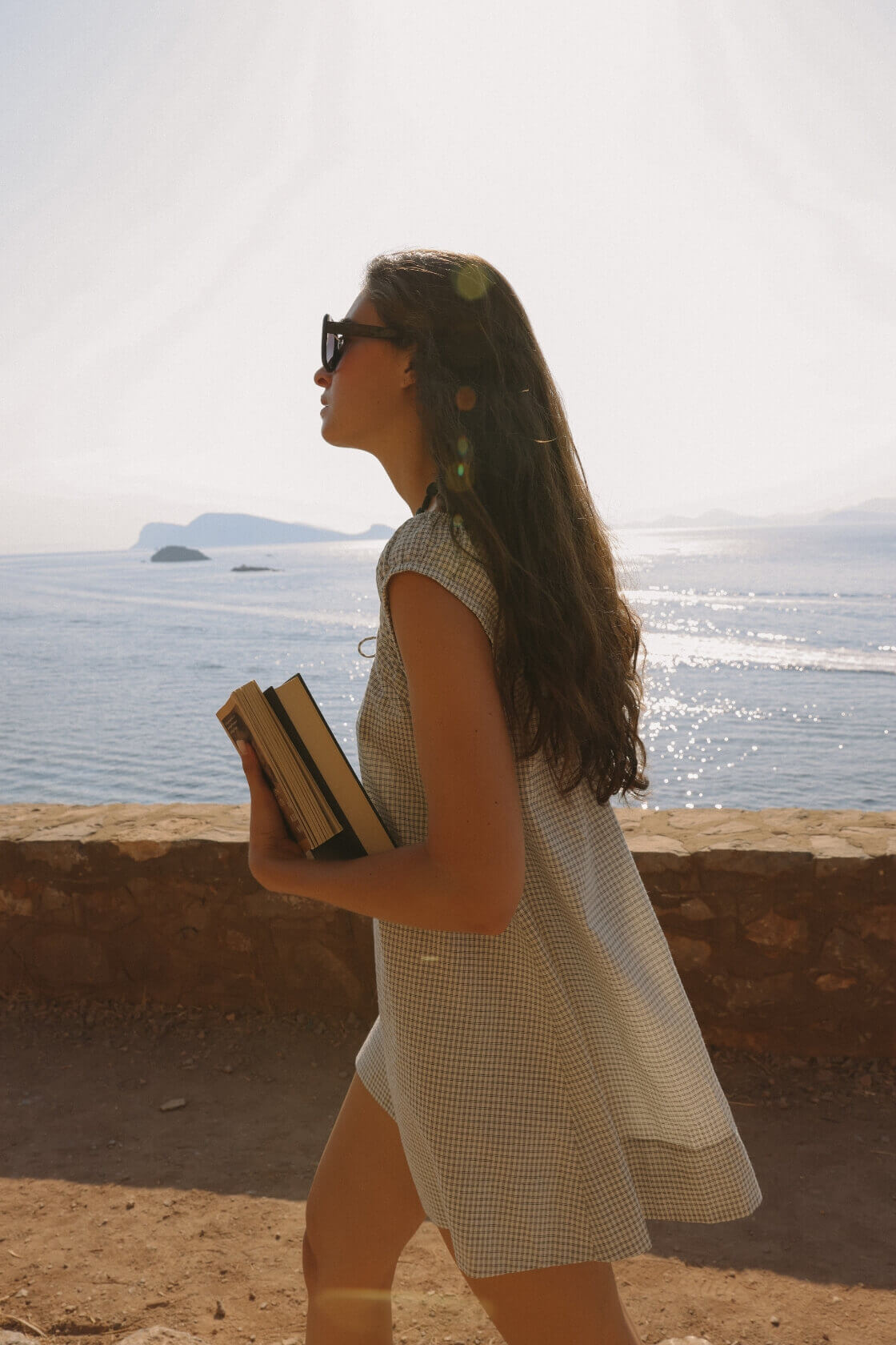 Model in a black check mini dress with flared silhouette and gathered neckline, walking seaside with books in hand—lightweight cotton ideal for warm days.