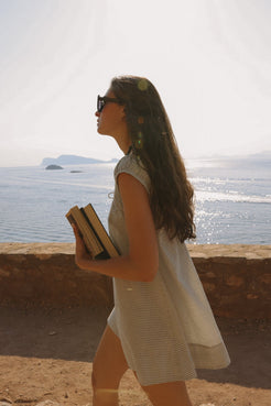 Model in a black check mini dress with flared silhouette and gathered neckline, walking seaside with books in hand—lightweight cotton ideal for warm days.