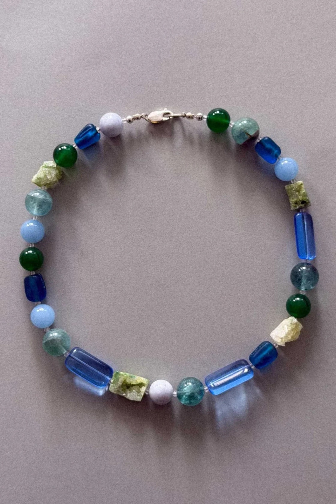 A beaded necklace with a mix of blue, green, and light blue translucent beads, some rectangular and some round, arranged in a circle on a neutral gray background.
