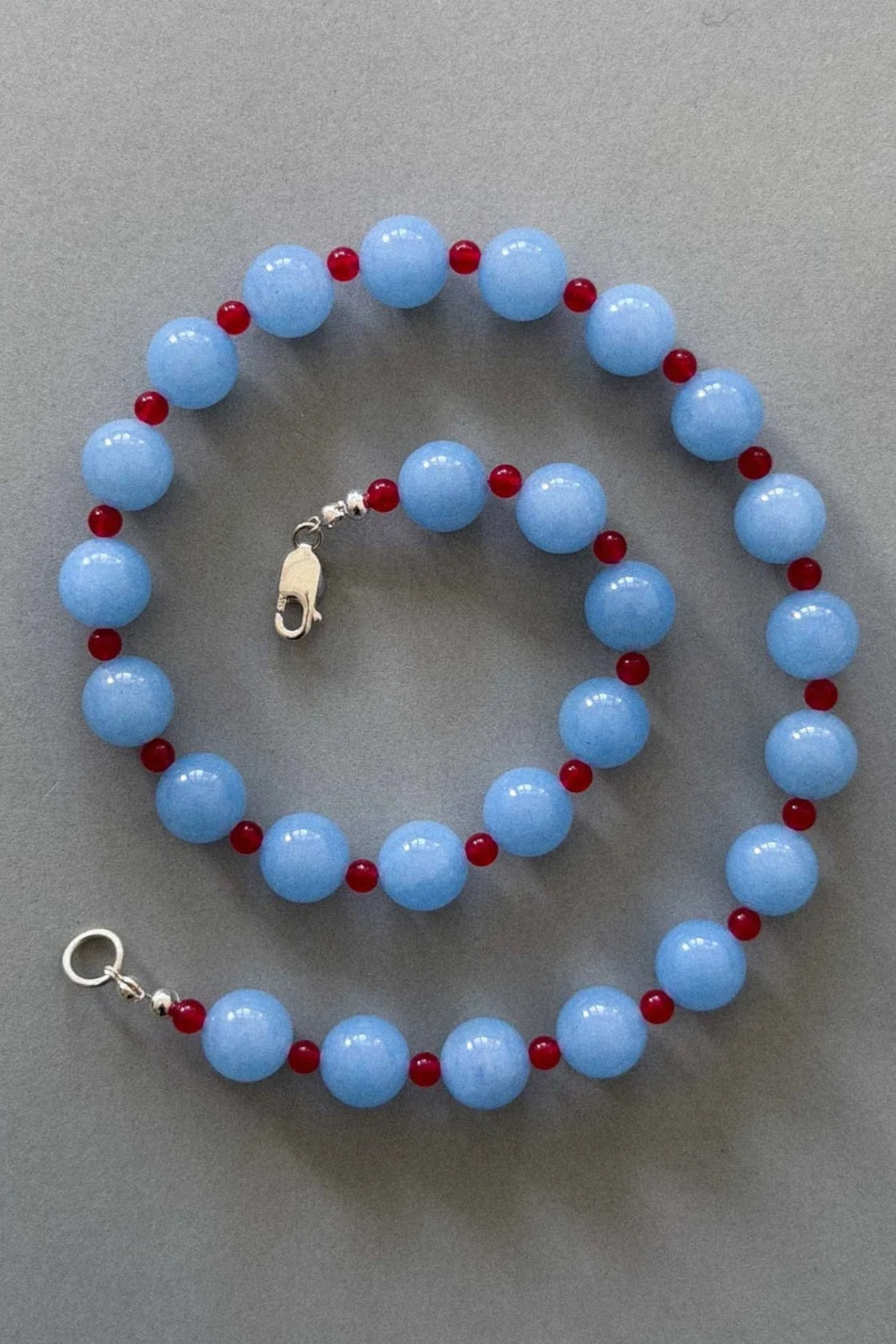 A beaded necklace and matching bracelet made of light blue round beads separated by small dark red beads, with silver-tone clasp hardware, displayed on a neutral gray background.