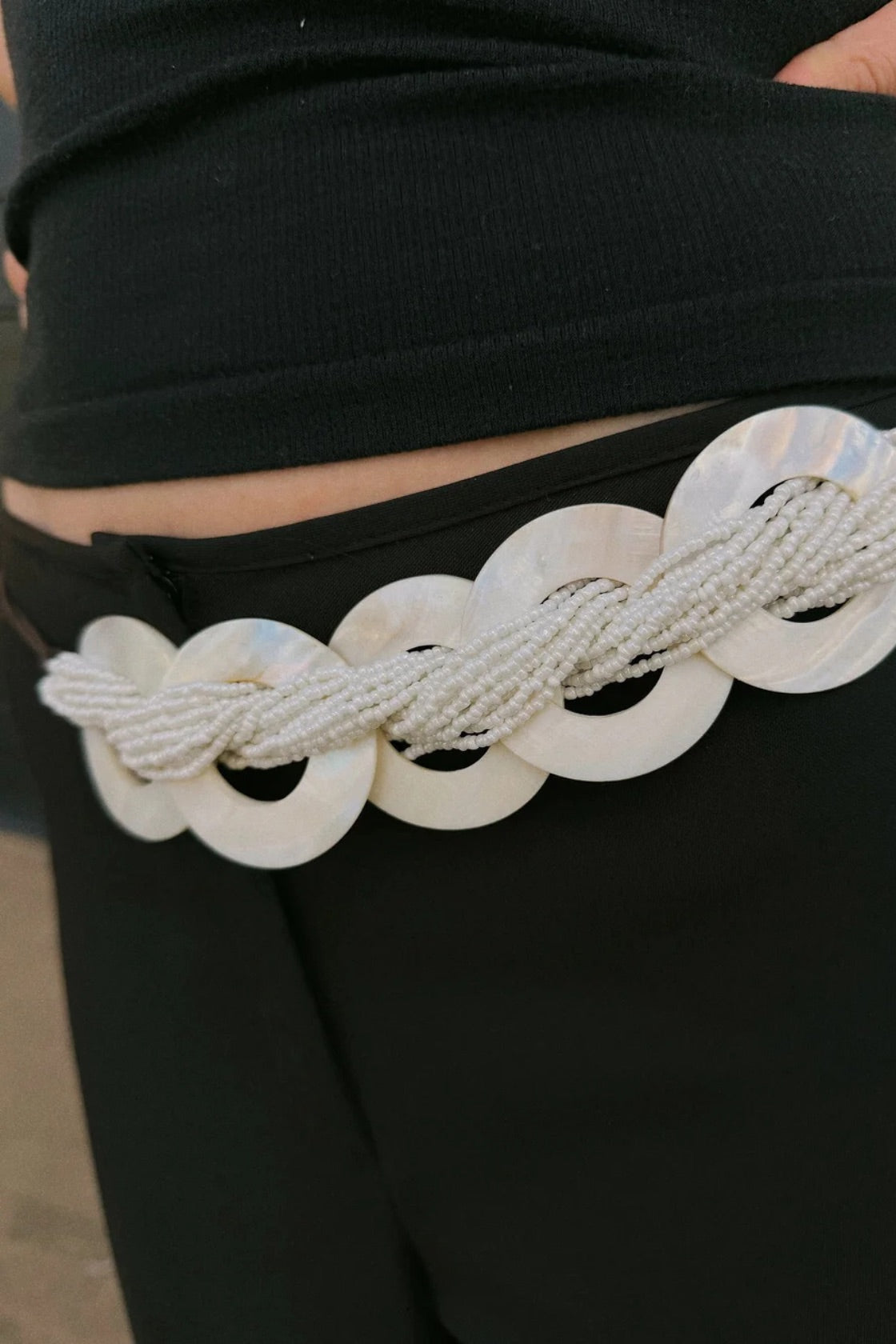A close-up of a person wearing a black ribbed top and black bottoms, featuring a decorative waist accessory with large white shell-like discs and a braided white rope belt.