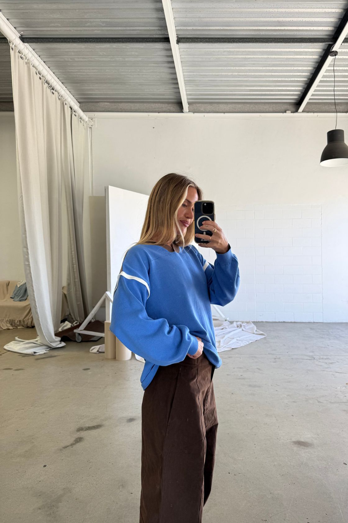 Mirror selfie lifestyle image of a model wearing a cobalt blue oversized sweatshirt with white stripe detail across the sleeves, relaxed fit and ribbed cuffs, paired with chocolate brown pants in a minimal studio space