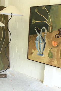 A framed still life painting hanging on a white brick wall, depicting a blue pitcher, green pear, and dark grapes on a tan and olive background. A black floor lamp with a white shade stands to the left in a room with a concrete floor.