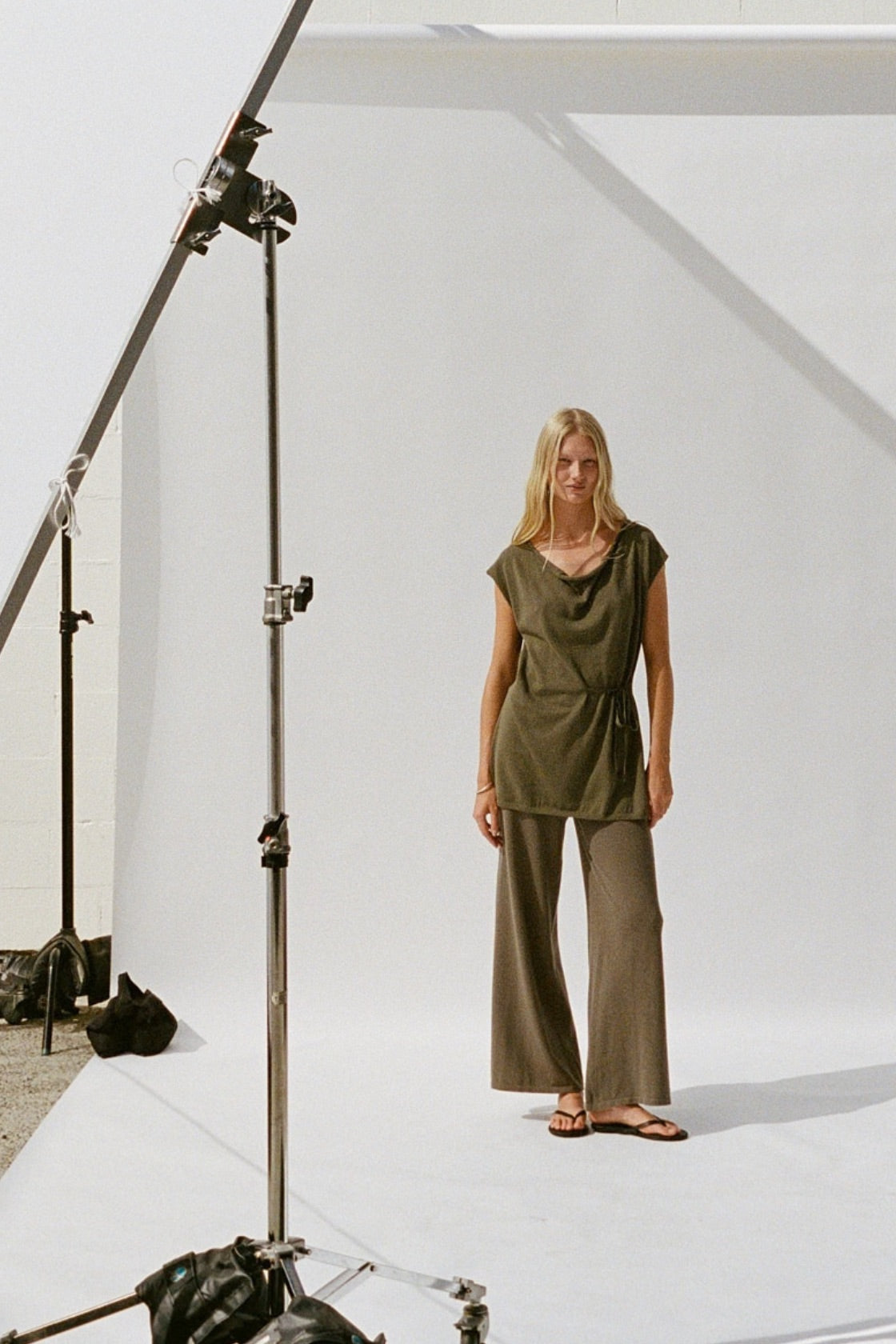 A woman stands on a white studio sweep wearing an olive green wrap-style top and matching wide-leg pants. Studio lighting equipment is visible on the left side of the frame.
