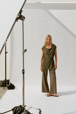 A woman stands on a white studio sweep wearing an olive green wrap-style top and matching wide-leg pants. Studio lighting equipment is visible on the left side of the frame.