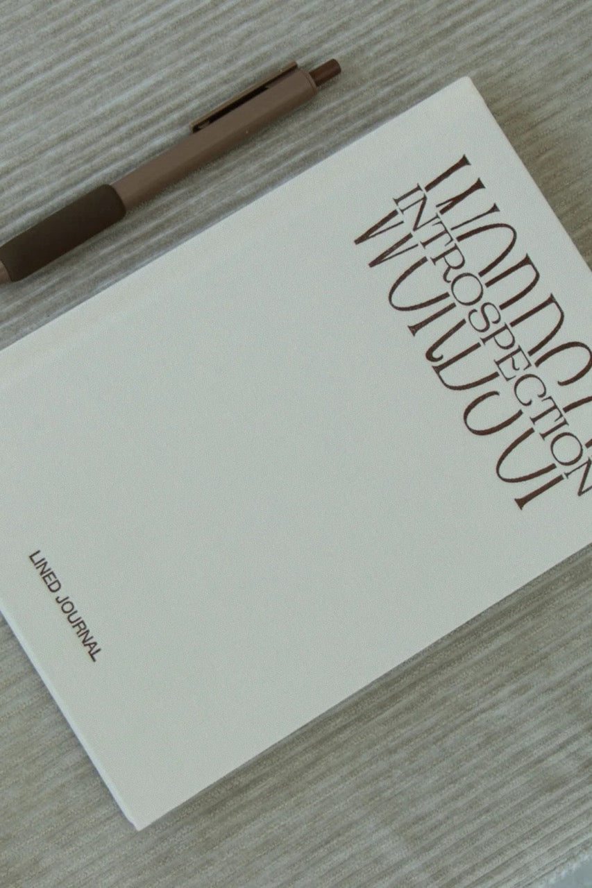 A lined journal with a minimalist off-white cover and large vertical title text reading 'Words of Introspection' lies on a light wood surface, with a brown pen placed diagonally across the top left.