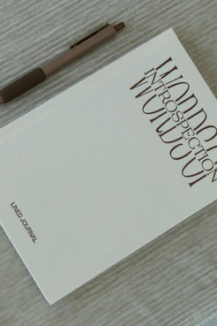 A lined journal with a minimalist off-white cover and large vertical title text reading 'Words of Introspection' lies on a light wood surface, with a brown pen placed diagonally across the top left.